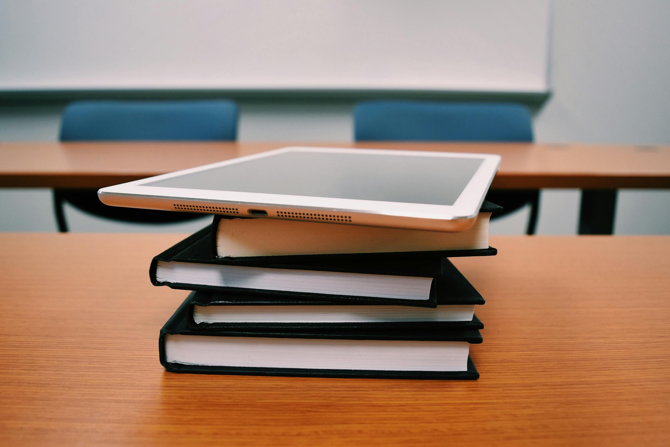 A tablet rests on top of a stack of books in an empty classroom, illustrating modern education.