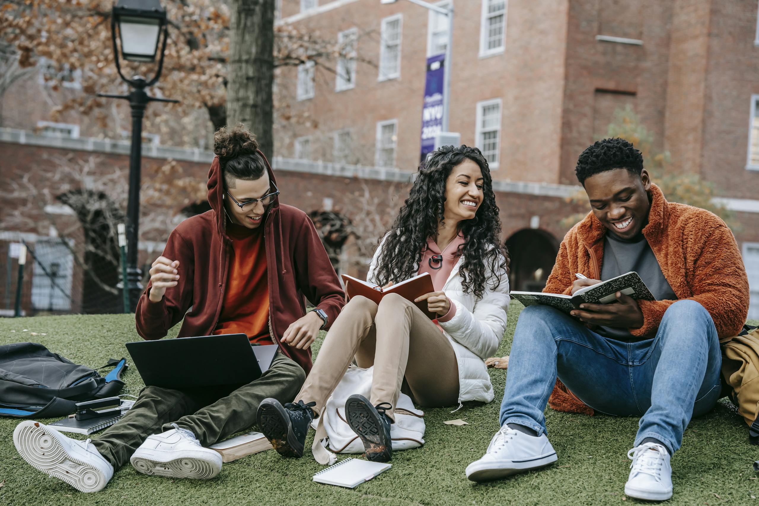 Full body of happy diverse students with notebooks and laptop sitting on grassy lawn on campus of university while studying together