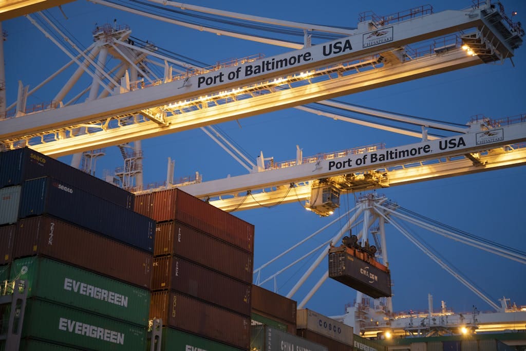 Illuminated cargo cranes at the Port of Baltimore with stacked containers at dusk.